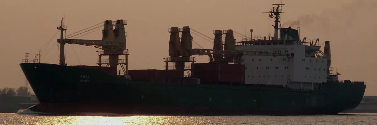 Silhouette of a general cargo ship with cranes at dusk, hazy orange sky and calm water