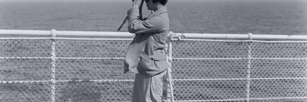 Black-and-white archive photo of a person at the ship's rail scanning the sea with binoculars